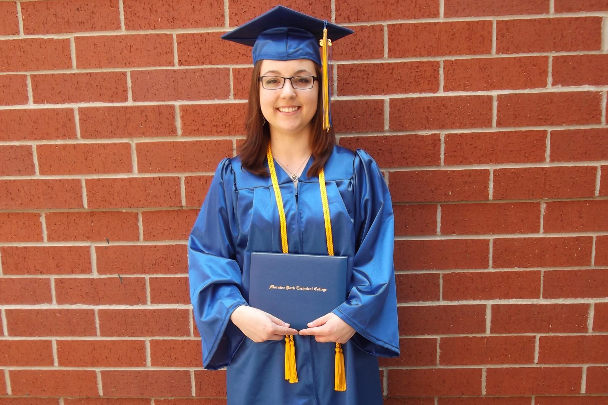 Female Moraine Park student standing against brick wall in grad apparel and diploma