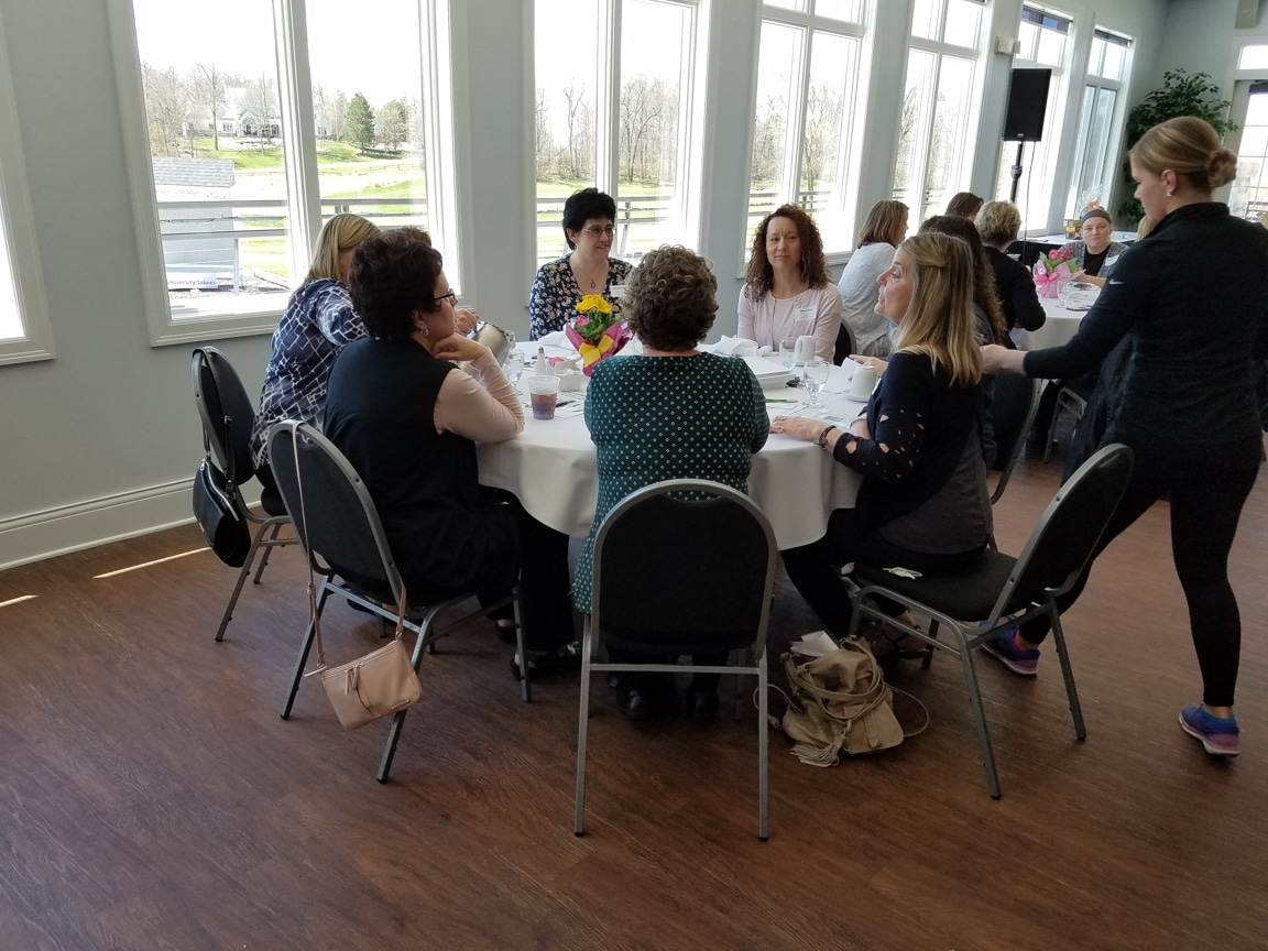 attendees sit around table at woman speaks at podium at mptc Wisconsin Women in Higher Education Leadership workshop