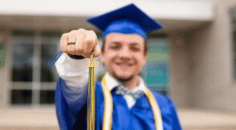 male student wearing blue graduation cap & gown, holding his tassel that says "2021"